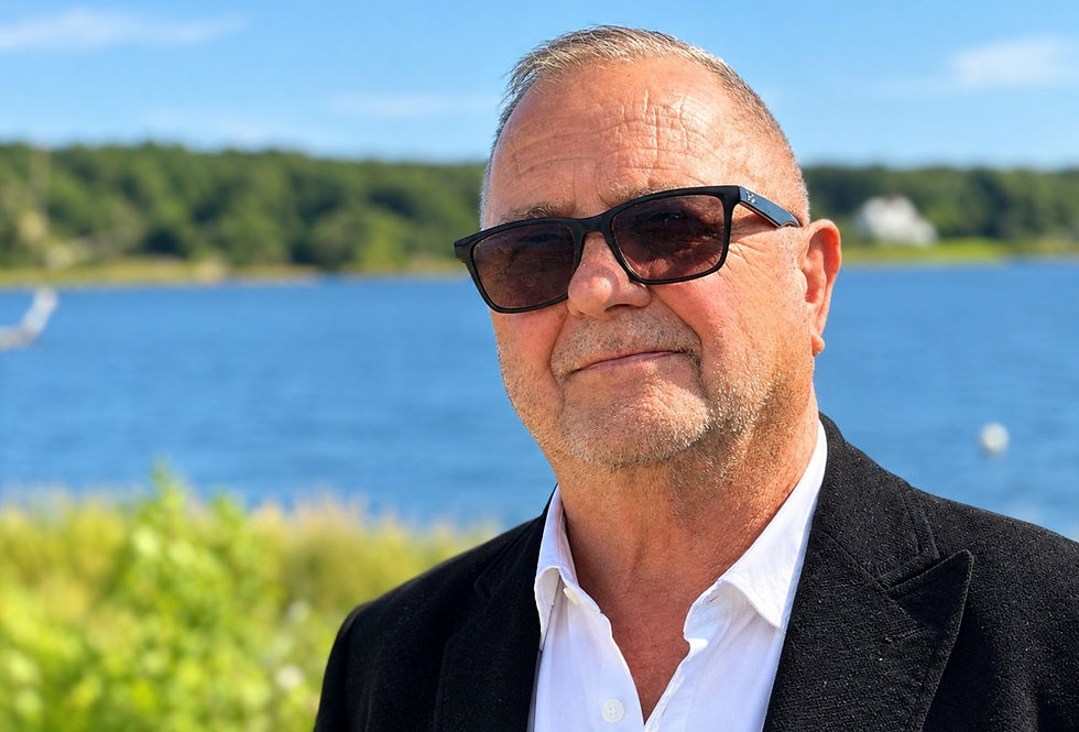 Older man in sunglasses and black blazer stands by a blue lake, looking calmly at the camera on a sunny day