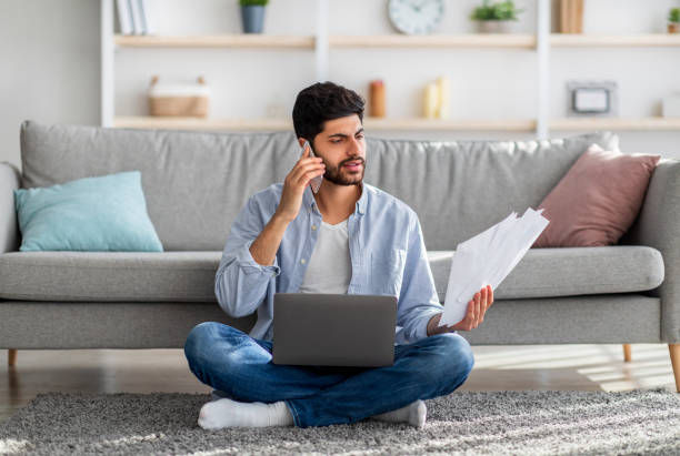 Man sitting cross-legged on a rug in a living room, talking on a phone while reviewing papers beside a laptop.