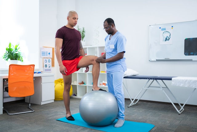 Physical therapist assists a man balancing on a gray exercise ball in a bright therapy room. Both focused, with medical diagrams in the background.