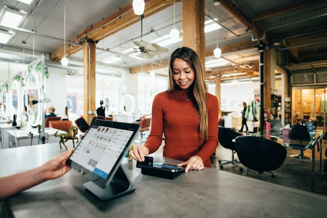 A smiling woman in a modern office or retail space making a payment at a point-of-sale terminal.