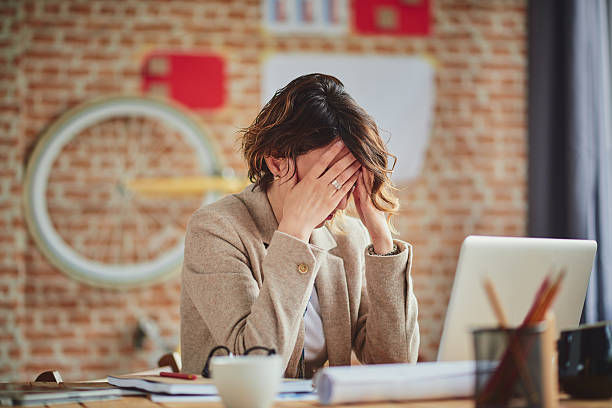 Woman in an office setting covers her face, appearing stressed. Background includes a brick wall and bicycle wheel. Papers and laptop on desk.