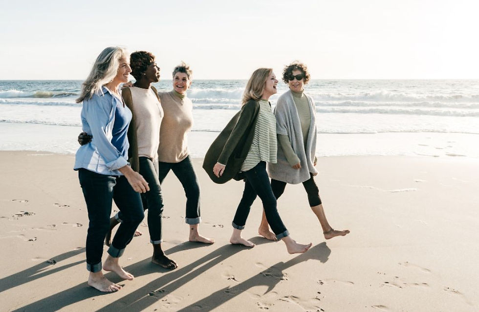 Five women walk along a sunny beach, smiling and barefoot. The ocean waves are in the background, creating a joyful and relaxed mood.