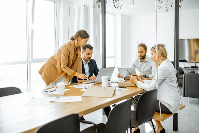 Four coworkers review laptops and a tablet around a conference table in a bright modern office.