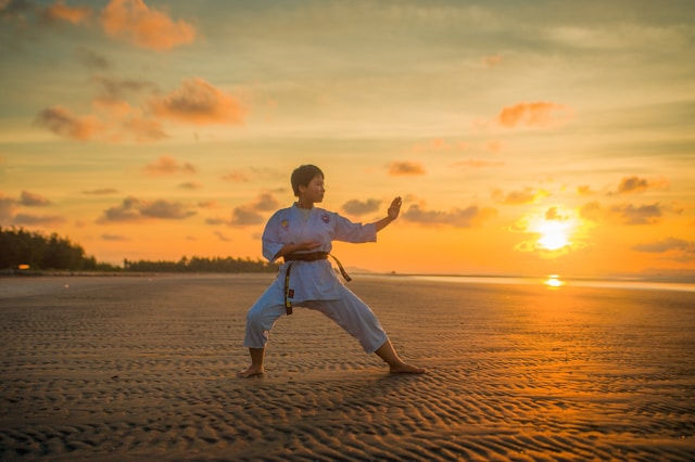 Martial artist in a white gi holds a karate stance on a beach at sunset, with golden sky and calm water behind.