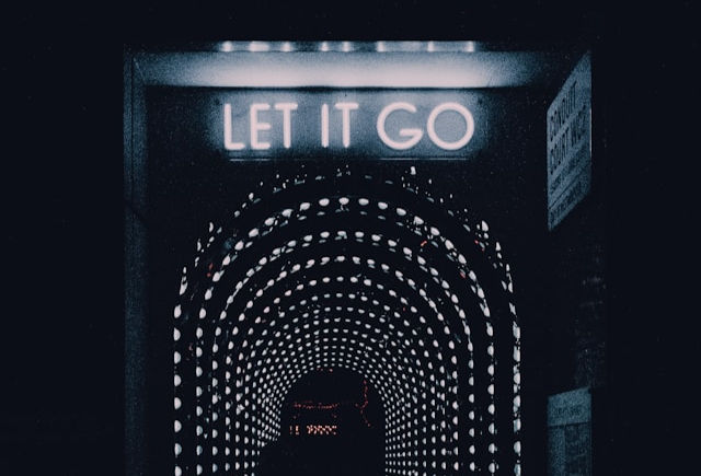 The image shows a dark, tunnel-like passage illuminated with dotted lights and a glowing neon sign at the top that reads "LET IT GO."