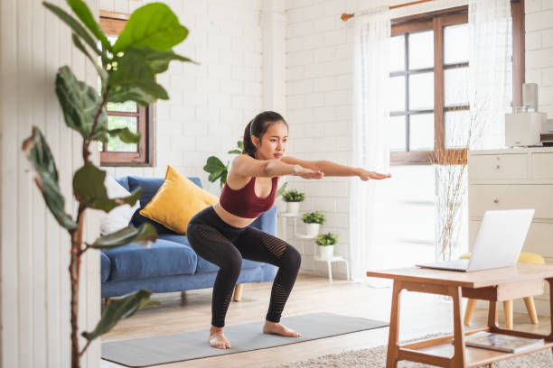 Woman in workout attire doing squats in a bright living room, with yoga mat, laptop, and plants. Blue sofa and yellow pillow in background.