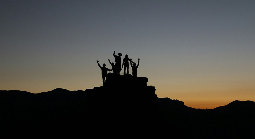 Silhouetted group of hikers cheering on a rocky peak at sunset, with orange sky and dark mountains above the horizon