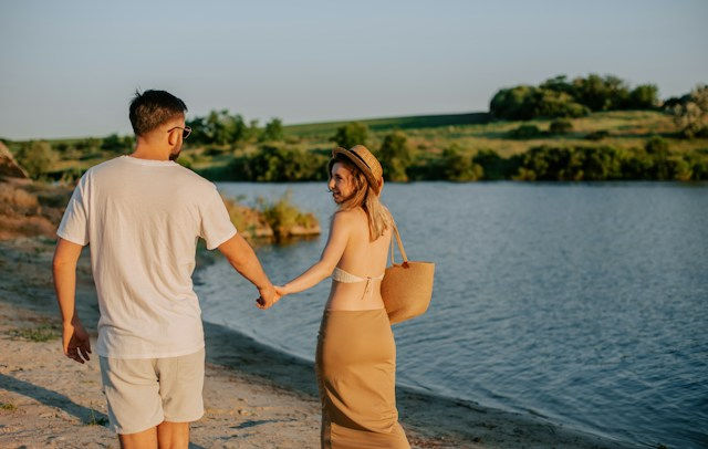 Couple holding hands by a lake at sunset, woman smiling in a hat and tan dress, with green hills in the background.