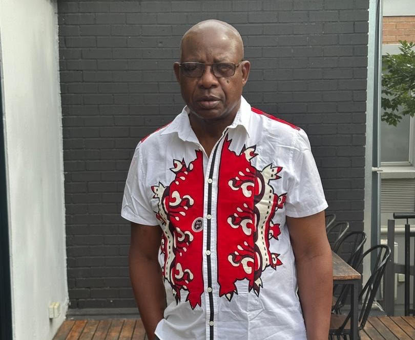 Man in a red and white patterned shirt stands outdoors against a dark brick wall. He appears calm, wearing glasses, with a subtle expression.