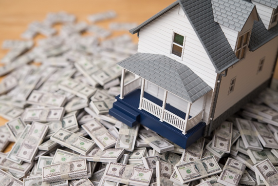 Model house on a pile of scattered dollar bills. House is white with gray roof. Background is a wooden surface. Mood suggests wealth.