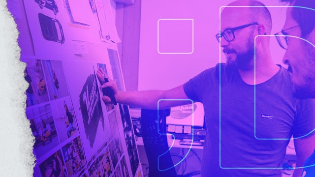 Man in glasses points at a wall of design sketches and photos in purple light, while another person watches nearby.