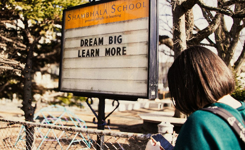Student with backpack reads a Shambhala School sign saying DREAM BIG LEARN MORE, outdoors among trees.