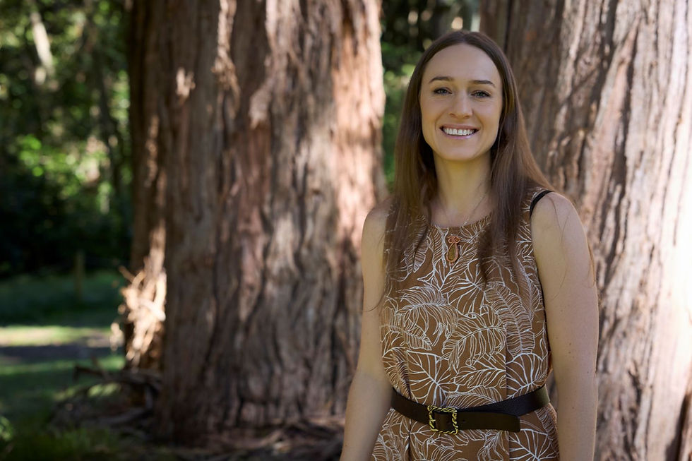 Smiling woman in a patterned dress stands by a giant tree in a sunny park.