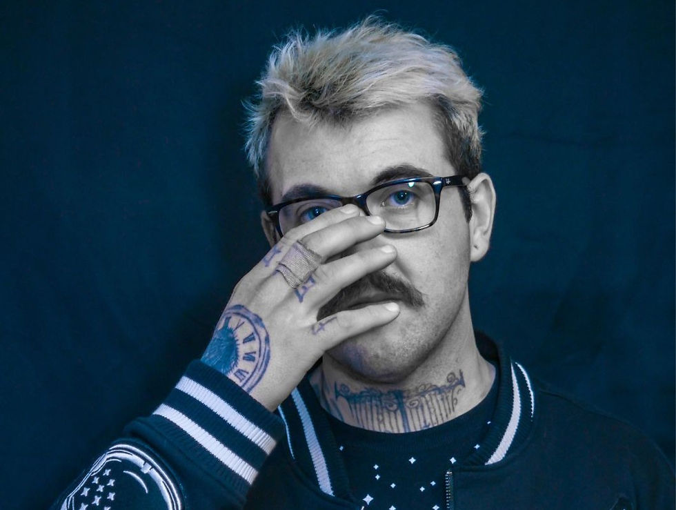 A person with blonde hair and glasses covers their mouth with a tattooed hand. Blue backdrop, wearing a black jacket with white stripes.