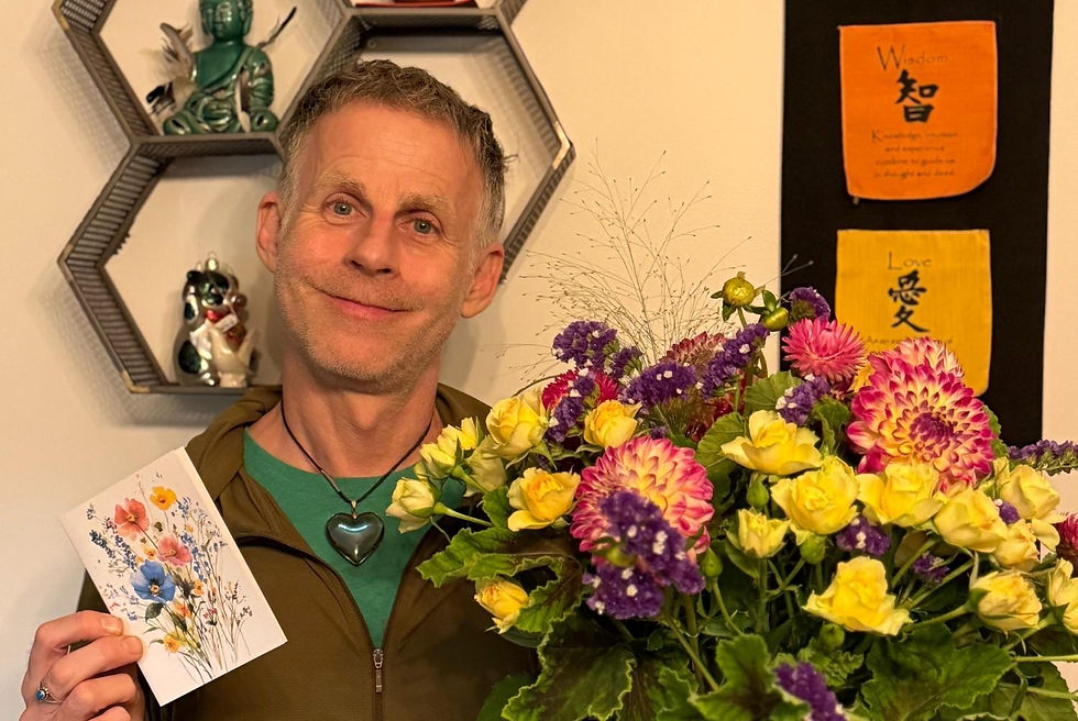 Smiling man holds a colorful flower bouquet and floral card in a cozy room with Buddha shelves and Wisdom/Love wall art.