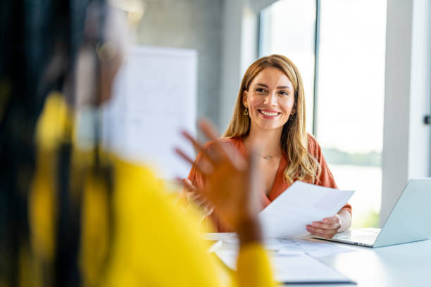 A smiling woman holding documents during a conversation or interview, sitting in a brightly lit office.