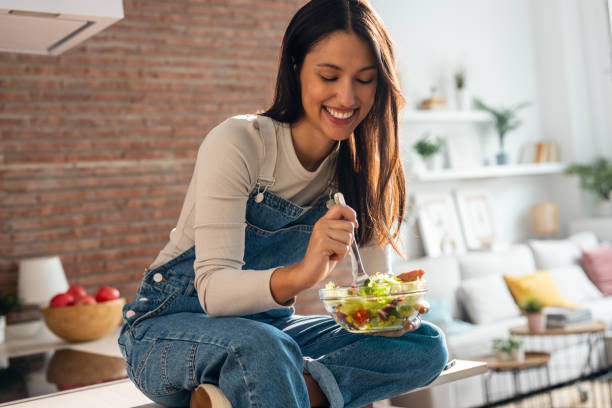 Woman in denim overalls sits on kitchen counter, smiling while eating salad from a glass bowl. Cozy living room with brick wall in background.