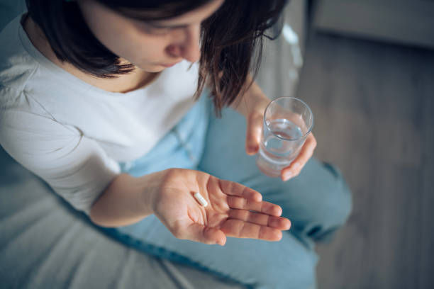 A person holds a pill in one hand and a glass of water in the other, sitting on a gray bed. The mood is contemplative.