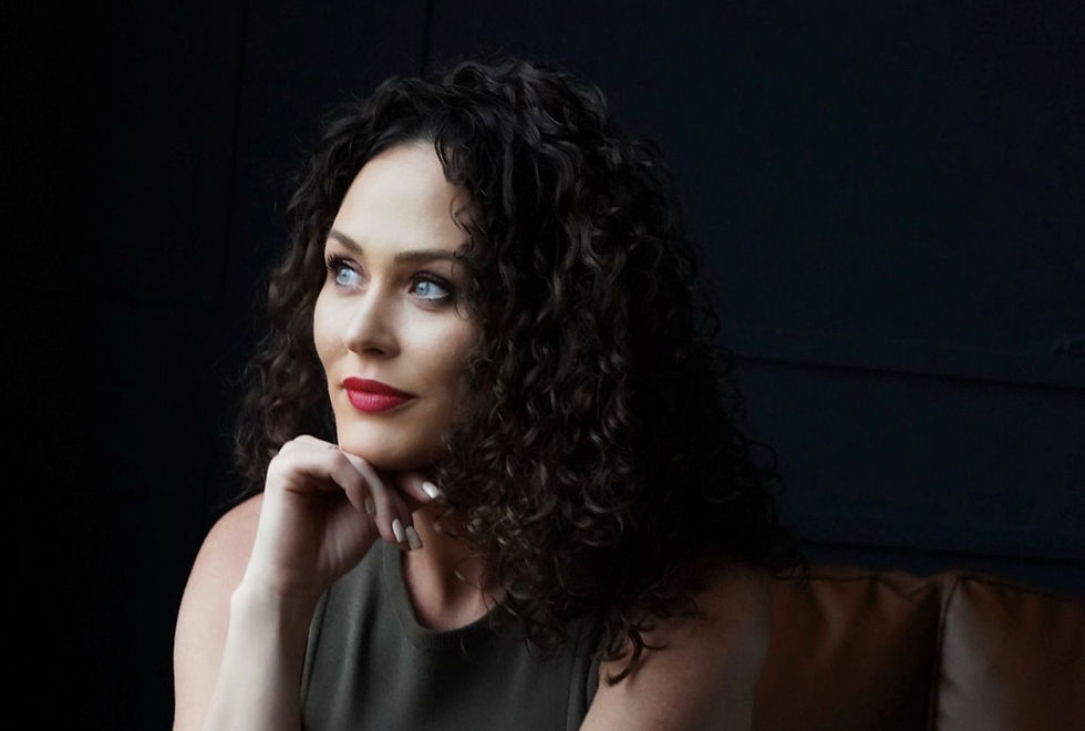 A woman with curly dark hair and red lipstick gazes thoughtfully to the side, resting her chin on her hand in a softly lit, dark-toned setting.