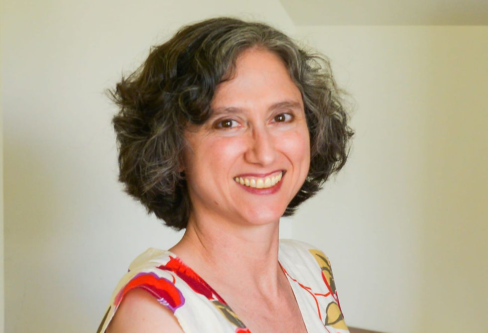 Smiling woman with short curly dark hair in a patterned white top, posing indoors against a plain light wall.
