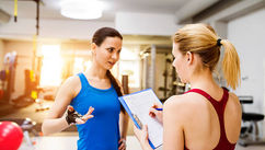 Two women in gym attire converse; one holds a clipboard. The bright gym setting features equipment. The mood appears focused and engaged.