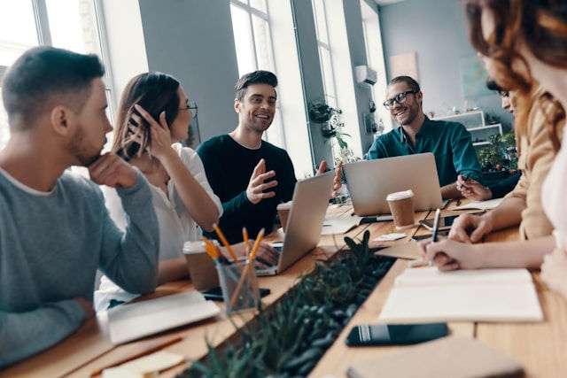 A group of young professionals is engaged in a lively discussion around a conference table, with laptops, notebooks, and coffee cups in front of them.