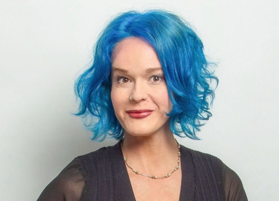 Woman with bright blue wavy hair smiles at camera against a white background, wearing a black top and necklace.