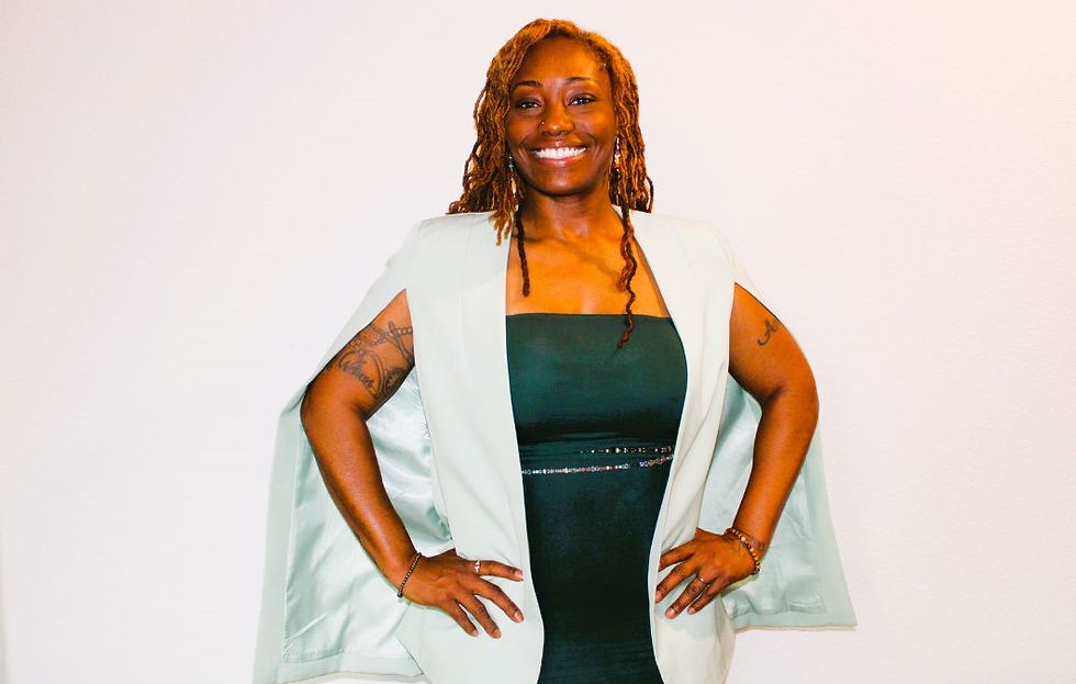 Smiling woman with dreadlocks poses confidently, wearing a green dress and light blazer. Tattoos visible on arms. Plain white background.