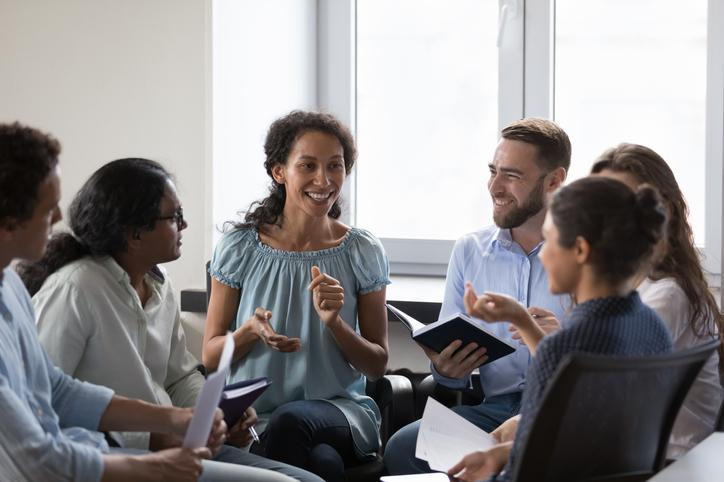 Smiling diverse group in a bright meeting room, seated in a circle and discussing with notebooks and papers in hand