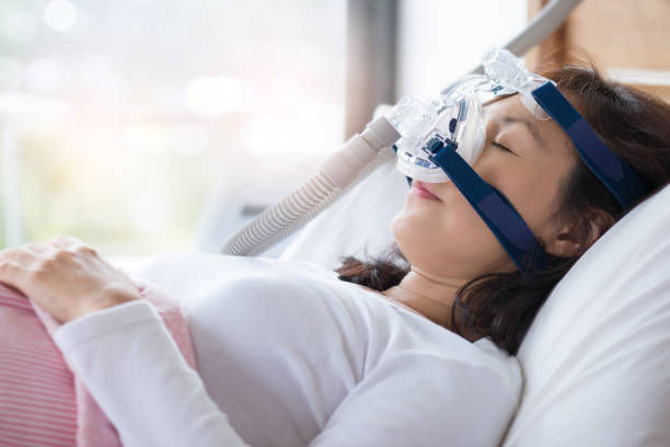 Woman asleep in bed wearing a CPAP mask for sleep apnea. Soft lighting from window, serene atmosphere, white bedding.