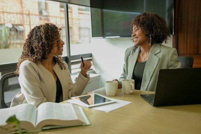 Two women smile in a bright office meeting, chatting over a laptop, mugs, tablet, and open book.