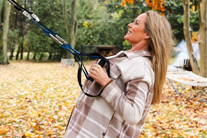 Woman in plaid coat holds blue handles, smiling in an autumn park with fallen leaves and trees in the background.