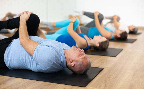 People lying on yoga mats in a studio, stretching on their backs. Bright clothing, wooden floor, calm and focused atmosphere.