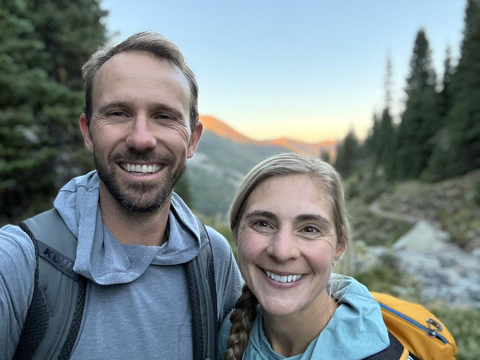 Smiling couple takes a hiking selfie in the mountains at sunset, with trees, backpacks, and glowing peaks.