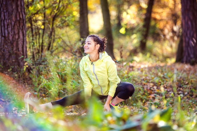 A woman in a yellow jacket stretches in a sunlit forest, with autumn leaves on the ground. She appears happy and relaxed.