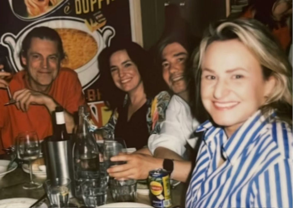 Four people smiling at a table with wine glasses and a soda can. Background shows part of a vintage advertisement. Warm, friendly atmosphere.