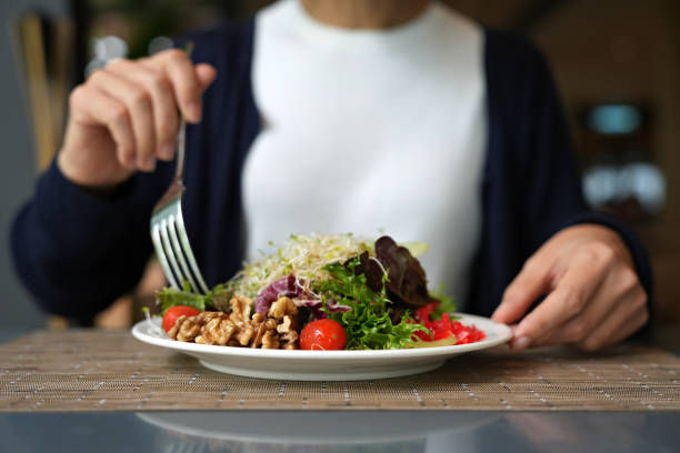 A person in a white shirt and dark sweater holds a fork, poised to eat a fresh salad with walnuts, tomatoes, and greens on a plate.