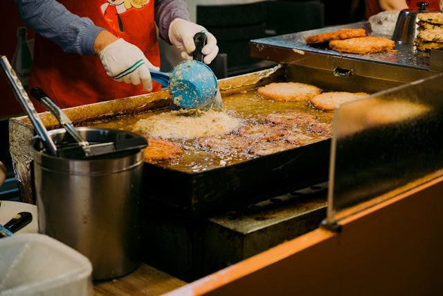 A person wearing gloves is deep-frying food, likely patties or cutlets, on a large griddle at a food stall or kitchen.