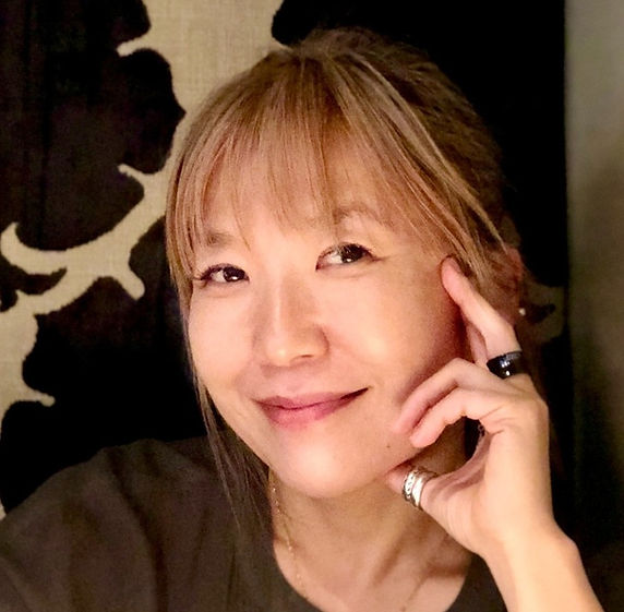 Smiling person with hand on face, wearing rings, against a dark patterned background. Cozy and warm ambiance.