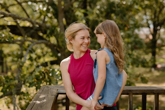 Smiling woman in a red top holds a girl in blue. They're on a wooden deck, surrounded by green trees, in a cheerful outdoor scene.