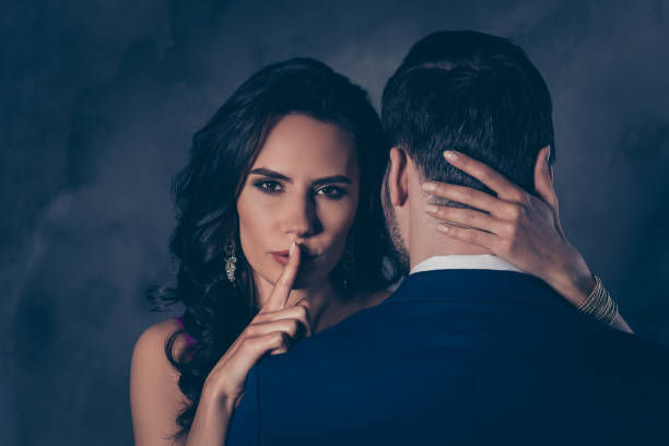 Woman with finger on lips signaling silence, embraces man in suit. Dark background, intimate mood. Her nails are light-colored.