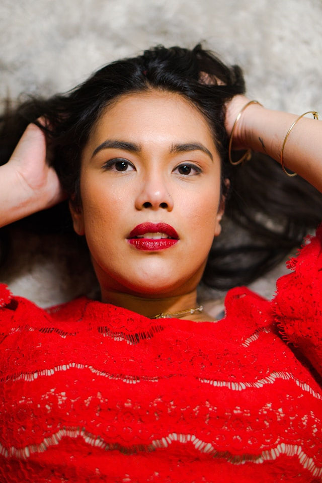 Woman in a red lace dress lies on a beige surface, hands behind head, with a calm expression. Bold red lipstick matches dress.