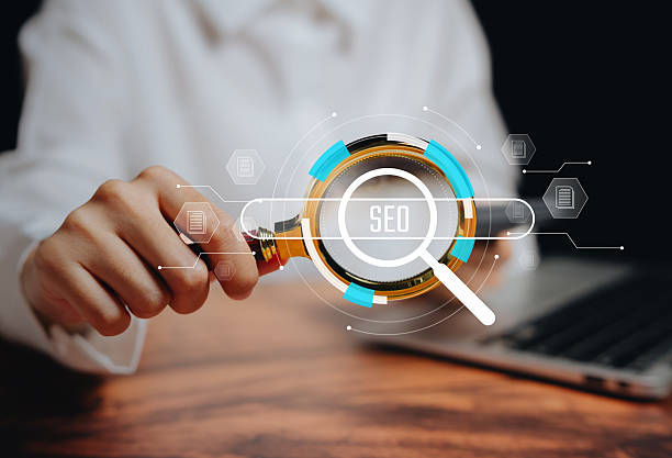 Person in white shirt holds a magnifying glass over a glowing SEO search graphic on a wooden desk beside a laptop.