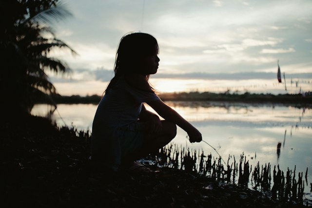 Silhouette of a child sitting by a calm lakeside at sunset, with palm trees and reflective water in the background.
