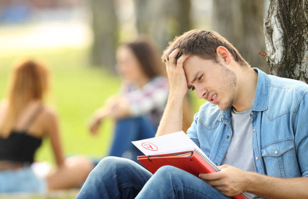 Male student staring on his failed exam paper while sitting on the grass on the school ground
