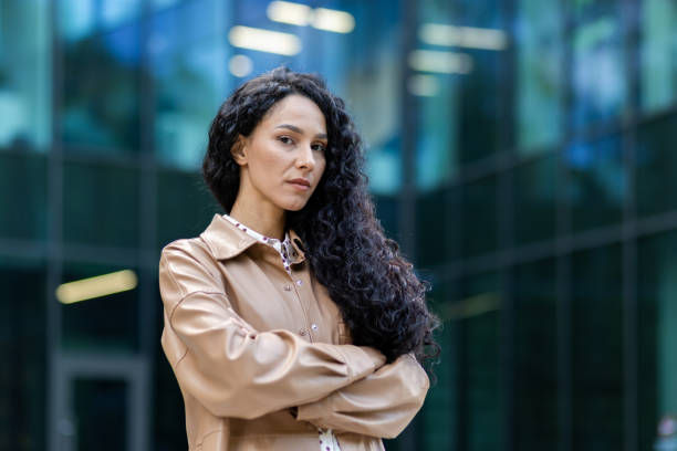 The photo shows a confident woman with long, curly black hair standing with her arms crossed in front of a modern glass building.