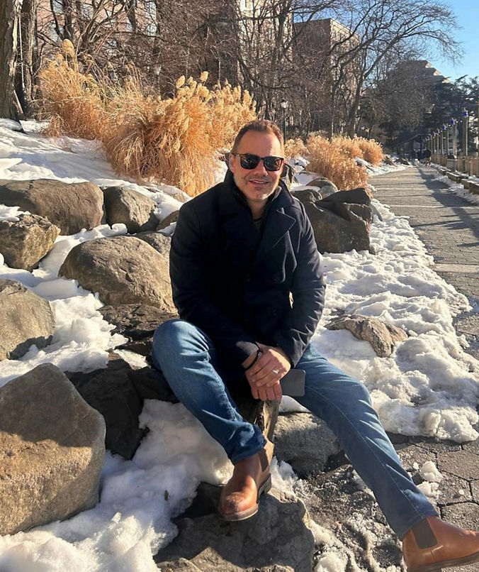 Man in sunglasses sits on a snowy rock in a park. He wears a dark coat and jeans. Sunlit grasses and bare trees in the background.