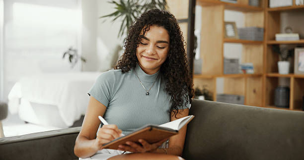 Smiling woman writes in a notebook on a couch in a bright living room with shelves and plants.
