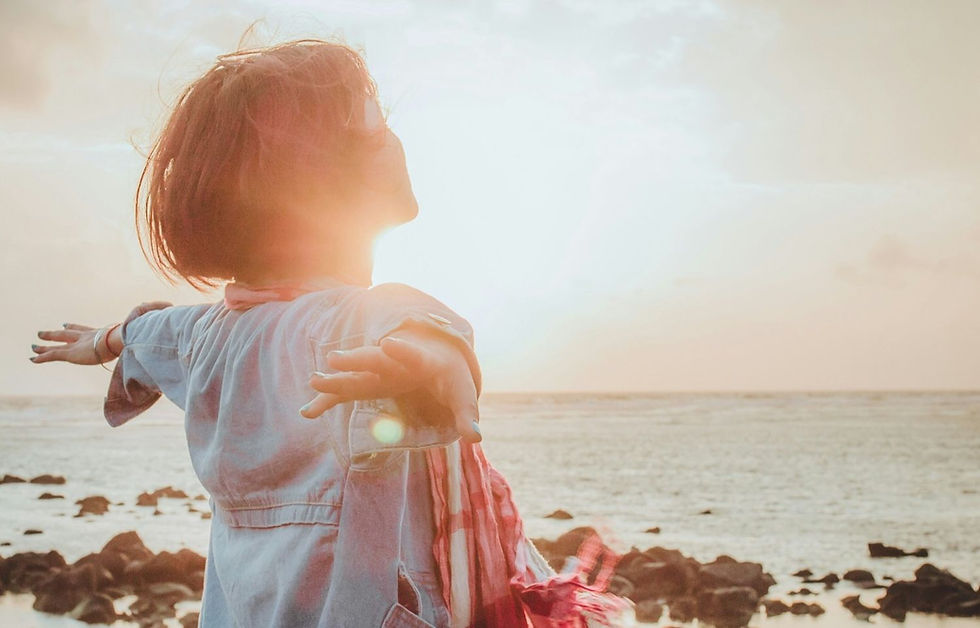 Backlit woman with outstretched arms on rocky beach, facing the sun and sea, scarf blowing in warm light.
