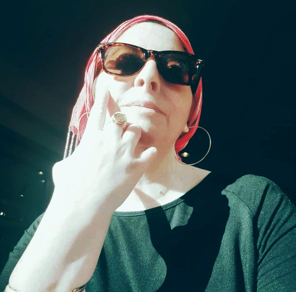 Woman in sunglasses and red headscarf sits in sunlight, wearing a ring and black top. She appears contemplative in a shaded room.